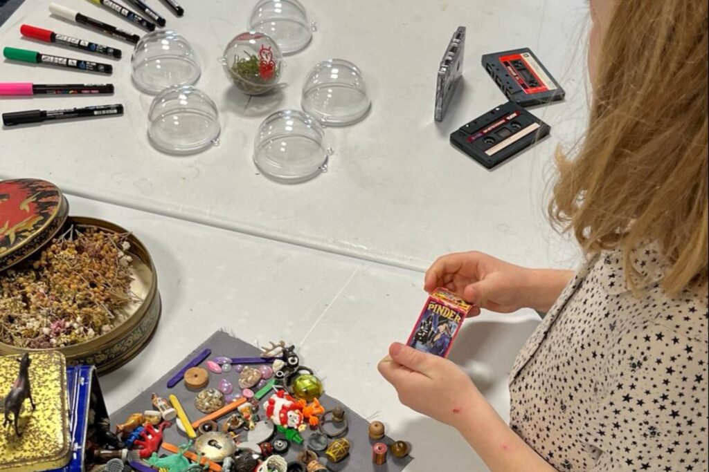 Une fillette à droite de l'image et devant une table blanche, manipule des jouets et figurines. Sur la tables sont posés des feutres, des cassettes audio, des sphères transparentes.