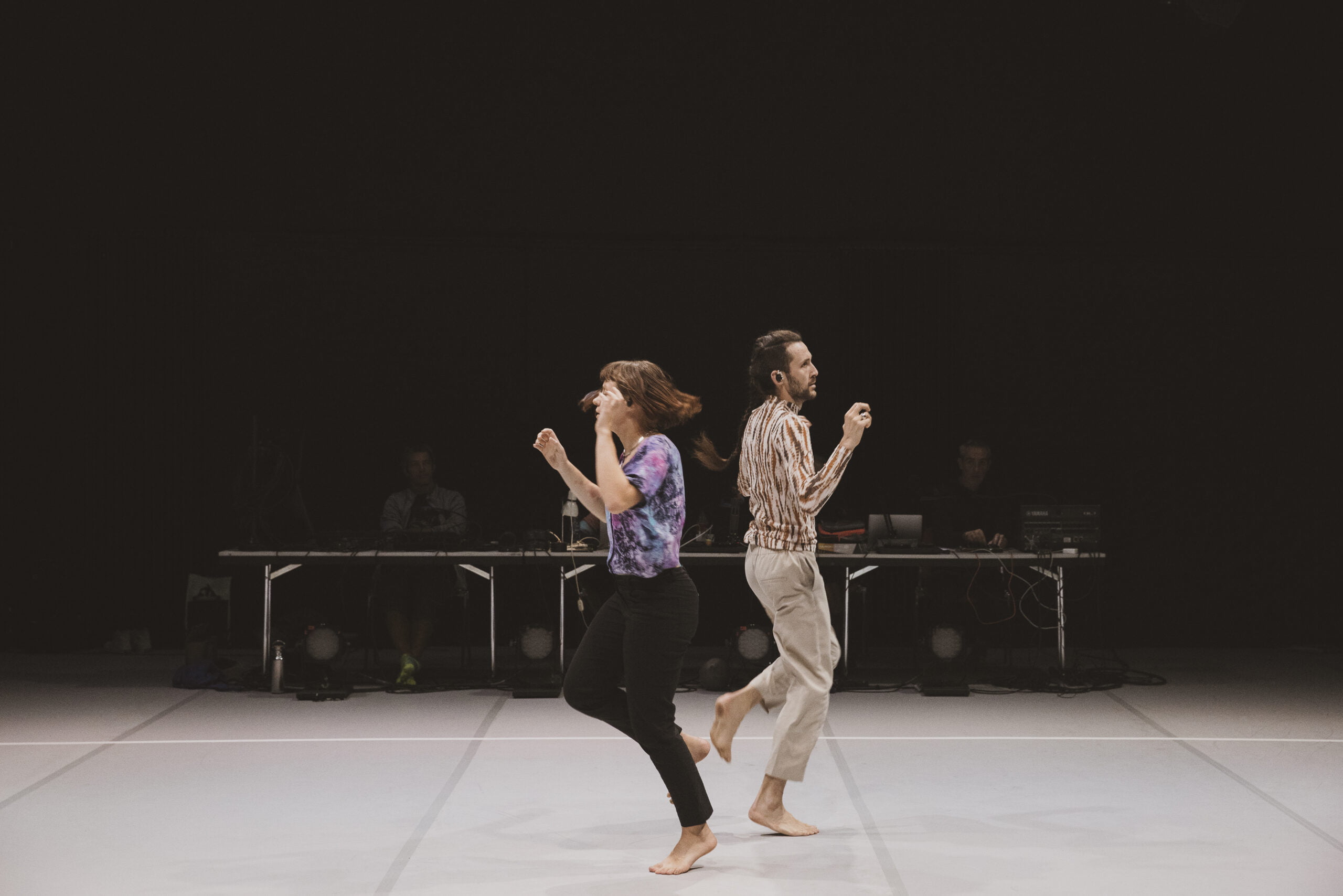 Sur un plateau de danse, Deux interprètes au centre de l'image, dansent dos à dos. A l'arrière plan on distingue la régie sur un fond noir.