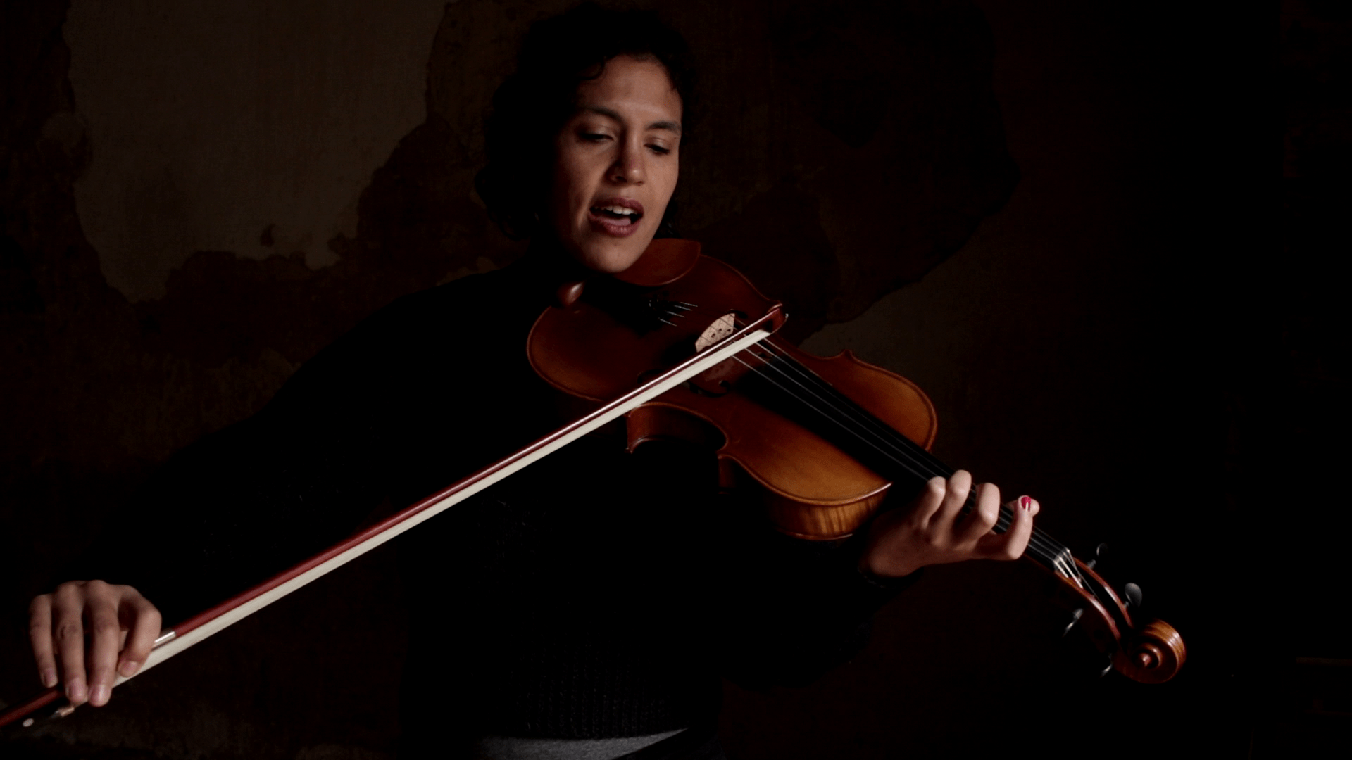 La violoniste Maud Herrera, brune, cheveux longs, visage doux et expressif, joue de son instrument face à l'objectif, dans une ambiance intimiste plongée dans la semi-pénombre,