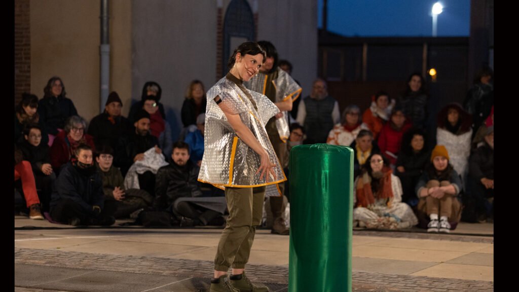 sur la place de la mairie, entourée de public la comédienne du collectif Le Thyase se tient debout, cheveux courts bruns lustrés, dans une pose gracieuse, elle porte un micro-cravate et un poncho transparent à pois jaunes, devant elle un pupitre fantaisiste en forme de boudin vert satiné