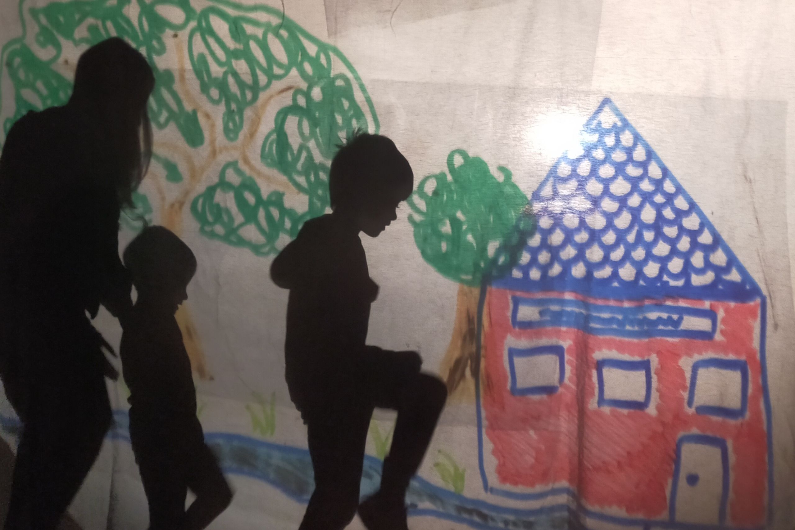 Dispositif de théâtre d'ombre grandeur nature : Un dessin d'enfant représentant un arbre et une maison sert de décor projeté. Trois silhouettes noir, un adulte et deux enfants viennent habiter se décor.