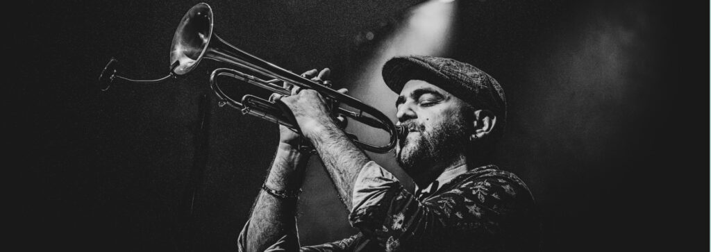 photo noir et blanc texturée où apparait le tropettiste de profil, casquette gavroche sur la tête, tenant son istrument de musique à hauteur de visage, yeux fermés sous le halo d'un projecteur de scène